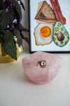 A light pink rose quartz bowl sits on a white table. There is a ring with a large green stone in the bowl. Behind the bowl is a green and purple plant in a gold pot on the left. On the right behind the bowl is a framed print called 'Boujee Brunch' 