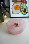 A light pink rose quartz bowl sits on a white table. There is a ring with a large green stone in the bowl. Behind the bowl is a green and purple plant in a gold pot on the left. On the right behind the bowl is a framed print called 'Boujee Brunch'