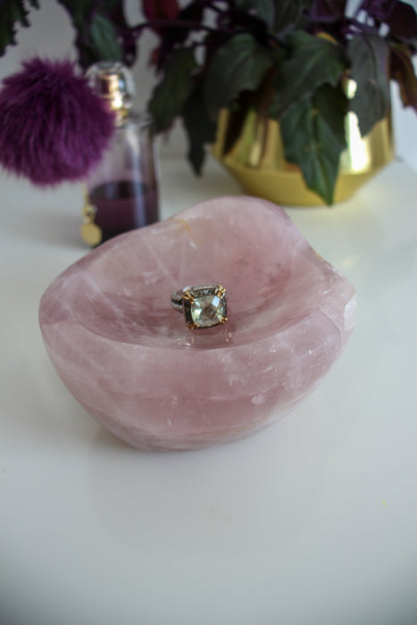 A dark pink rose quartz bowl sits on a white table. On the right behind the rose quartz bowl is a green and purple plant in a gold pot. On the left, behind the rose quartz bowl is a bottle of perfume, with a purple pom pom and it contains purple liquid. In the rose quartz bowl sits a fancy cocktail ring with a huge pale green faceted stone.