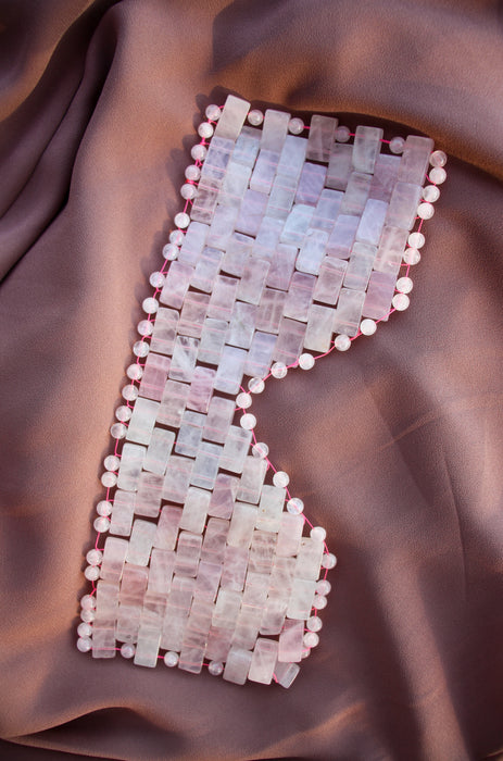 A light pink Rose Quartz eye mask sits on top a dusky pink background. The Rose Quartz eye mask is comprised of multiple small tiles of crystal and bound together with pink thread. 