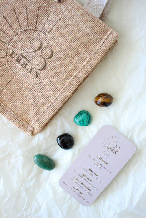 This image shows the 23 Urban Taurus Zodiac Kit in flat lay. To the upper leeft of the picture is the 23 Urban jute bag which all products come in. Then are the four Taurus tumble stones, which are brown and yellow Tigers Eye, green Malachite, Black Tourmaline and green Aventurine. Next to the tumble stones on the bottom right is the Taurus Zodiac Crystal Kit card, outlining the traits of Taurus and explaining the properties of the crystals.  