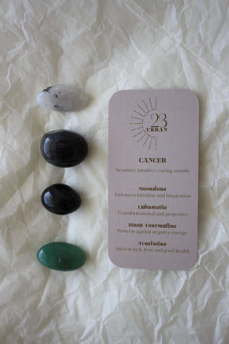 To the left of this image are the four Cancer Zodiac Crystal Kit tumble stones, which are Moonstone, Labradorite, Black Tourmaline and green Aventurine. Next to the tumble stones is a Cancer Zodiac Crystal Kit card, outlining the traits of Cancer and explaining the properties of the crystals. 