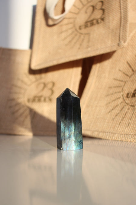 A Labradorite Tower crystal with an intense blue flash stands on a white table. In the background are 23 Urban branded jute bags. 