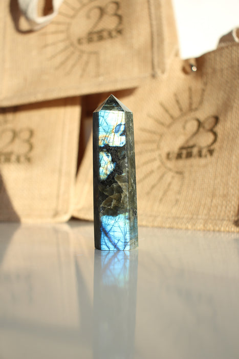 A Labradorite Tower crystal with an intense blue flash stands on a white table. In the background are 23 Urban branded jute bags. 