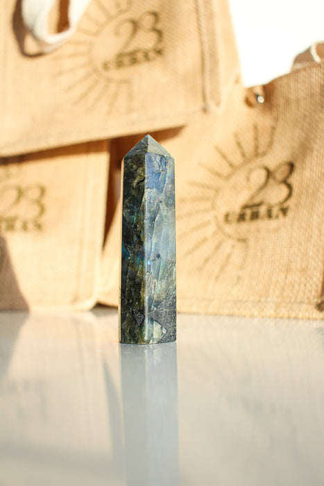 A Labradorite Tower crystal with an intense blue flash stands on a white table. In the background are 23 Urban branded jute bags. 