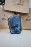 A Labradorite Freeform crystal with an intense blue flash stands on a white table. In the background are 23 Urban branded jute bags. 