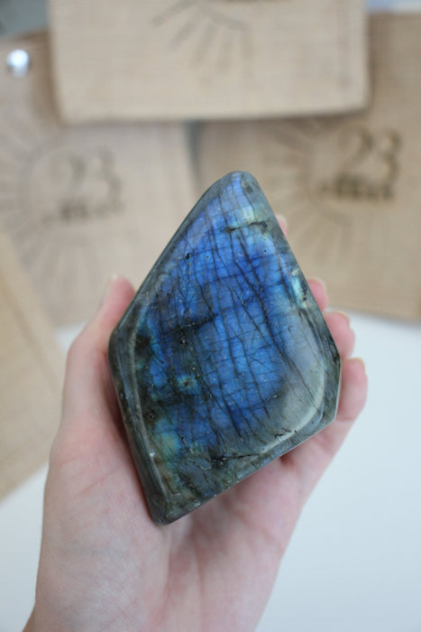 A Labradorite Freeform crystal with an intense blue flash stands on a white table. In the background are 23 Urban branded jute bags. 