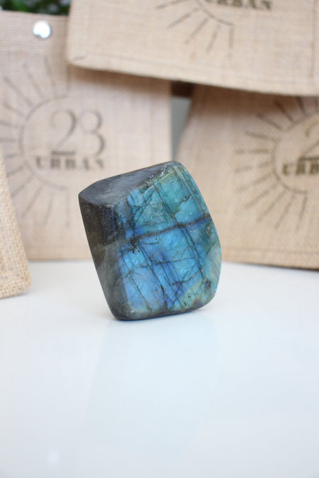 A Labradorite Freeform crystal with an intense blue flash stands on a white table. In the background are 23 Urban branded jute bags. 