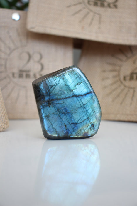 A Labradorite Freeform crystal with an intense blue flash stands on a white table. In the background are 23 Urban branded jute bags. 