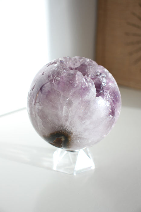 A beautiful Uruguayan Amethyst Sphere sits on a white bookshelf in the afternoon sun 