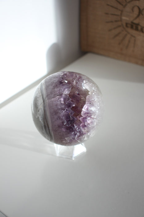 A juicy purple and white amethyst sphere sits on a white bookshelf in the afternoon sunlight