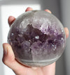 A juicy purple and white amethyst sphere sits on a white bookshelf in the afternoon sunlight