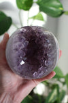 A large juicy purple Amethyst Sphere is held up by a hand in the afternoon sunlight