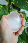 A glowing white selenite palm stone sits in a persons hand. in the background is a green plant