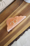 A peach coloured, cloud shaped cleansing plate sits flat on a brown table. On it sits two gold rings