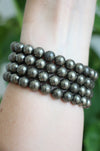 A deep golden stack of Pyrite bracelets on a wrist in front of a green plant 