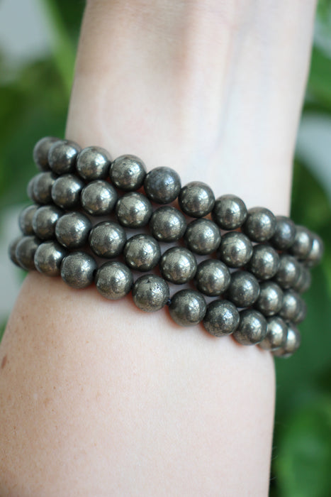 A deep golden stack of Pyrite bracelets on a wrist in front of a green plant 